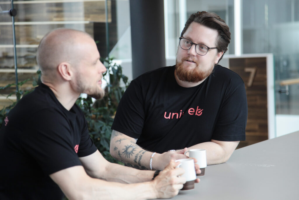 To Uniweb-konsulenter i samtale rundt et møtebord, begge med Uniweb-logotrykk på svarte t-skjorter. De diskuterer profesjonelt mens de holder kaffekopper, i et moderne kontormiljø med planter og glassvegger som symboliserer åpen kommunikasjon.