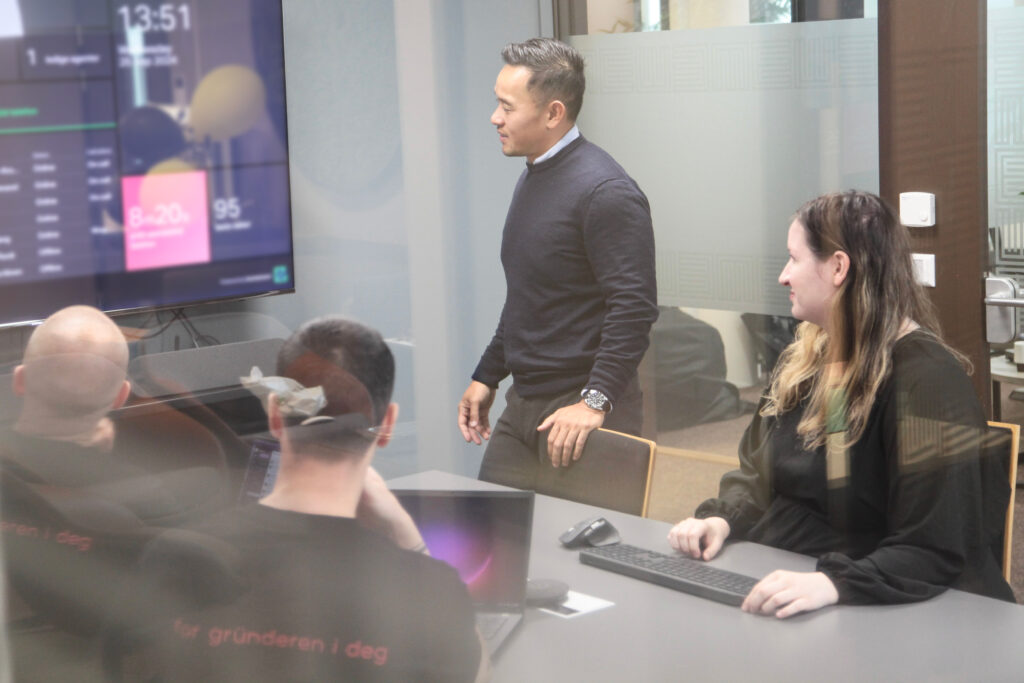 En Uniweb-ansatt står ved et møtebord og forklarer mens kollegaer sitter rundt bordet med datamaskiner